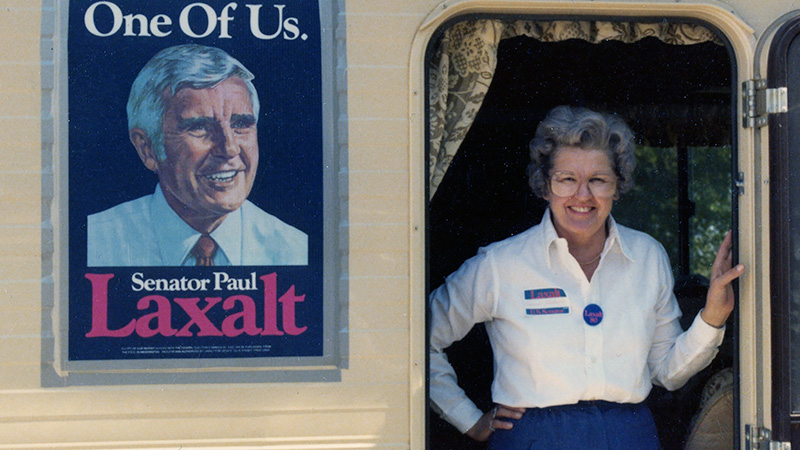 Barbara Vucanovich in a white blouse and blue skirt stands in the doorway of a camper van, with a poster on the side promoting Paul Laxalt. Barbara F. Vucanovich Papers, 96-54_10_146.