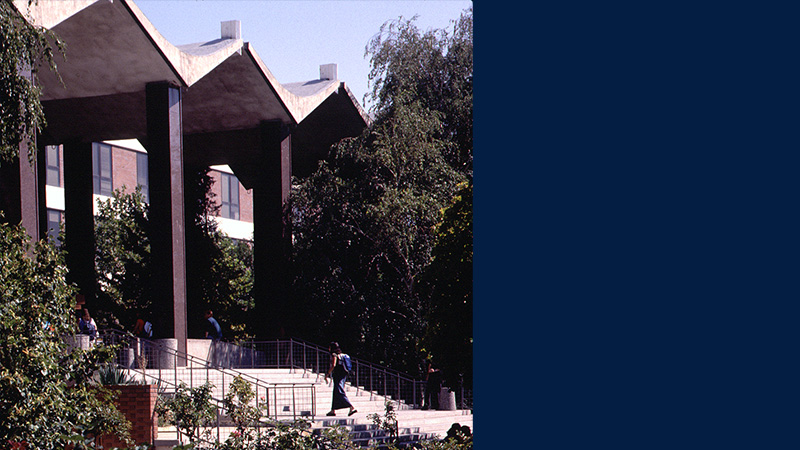 Red roses are in full bloom outside the Getchell Library with students walking on the library steps in the background. ID: UNRA-P3600-05837.