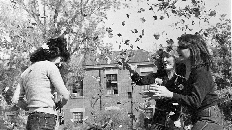 Female students laugh as they throw fall leaves at each other near Manzanita Lake with Clark Administration Building in the background.
