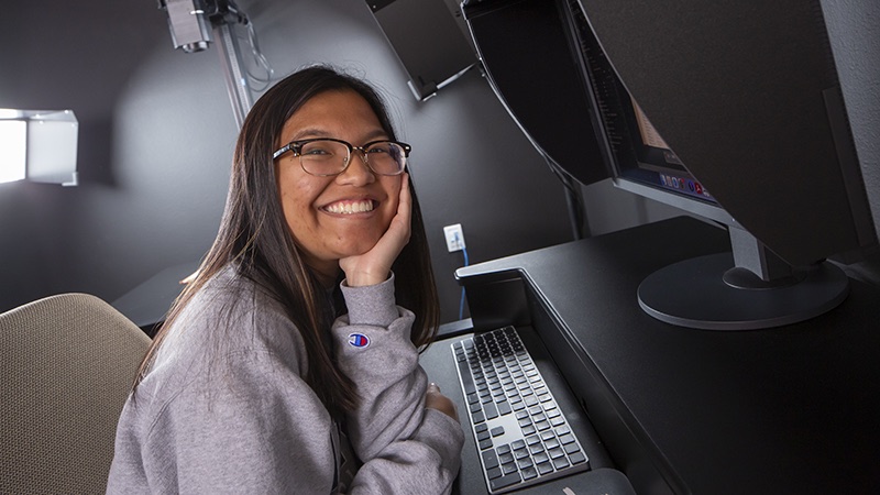A student assistant smiles while seated at a computer station.