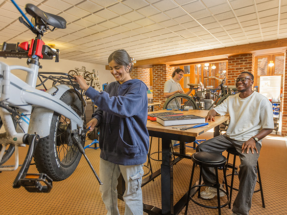Two people work on bicycles in a bike repair lab while another person sits at a workbench nearby.