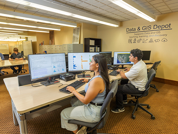 Two people work at computer stations in the Data & GIS Depot, with data visualizations and maps displayed on multiple monitors.