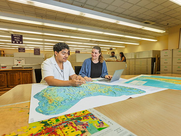 Two people examine a large map spread across a table, with additional maps and a laptop nearby.