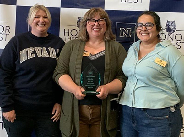 Three women stand smiling. The middle woman, Molly Mott, holds an award.