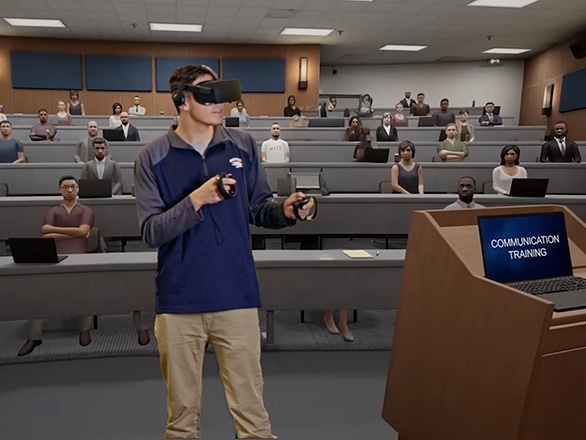 A person stands with a virtual reality headset on and holds controls. A virtual classroom is in the background with a laptop on a podium that reads " Communication training".