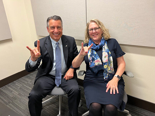 President Brian Sandoval and Dean Cardwell sit together making Wolfpack symbols with their hands.