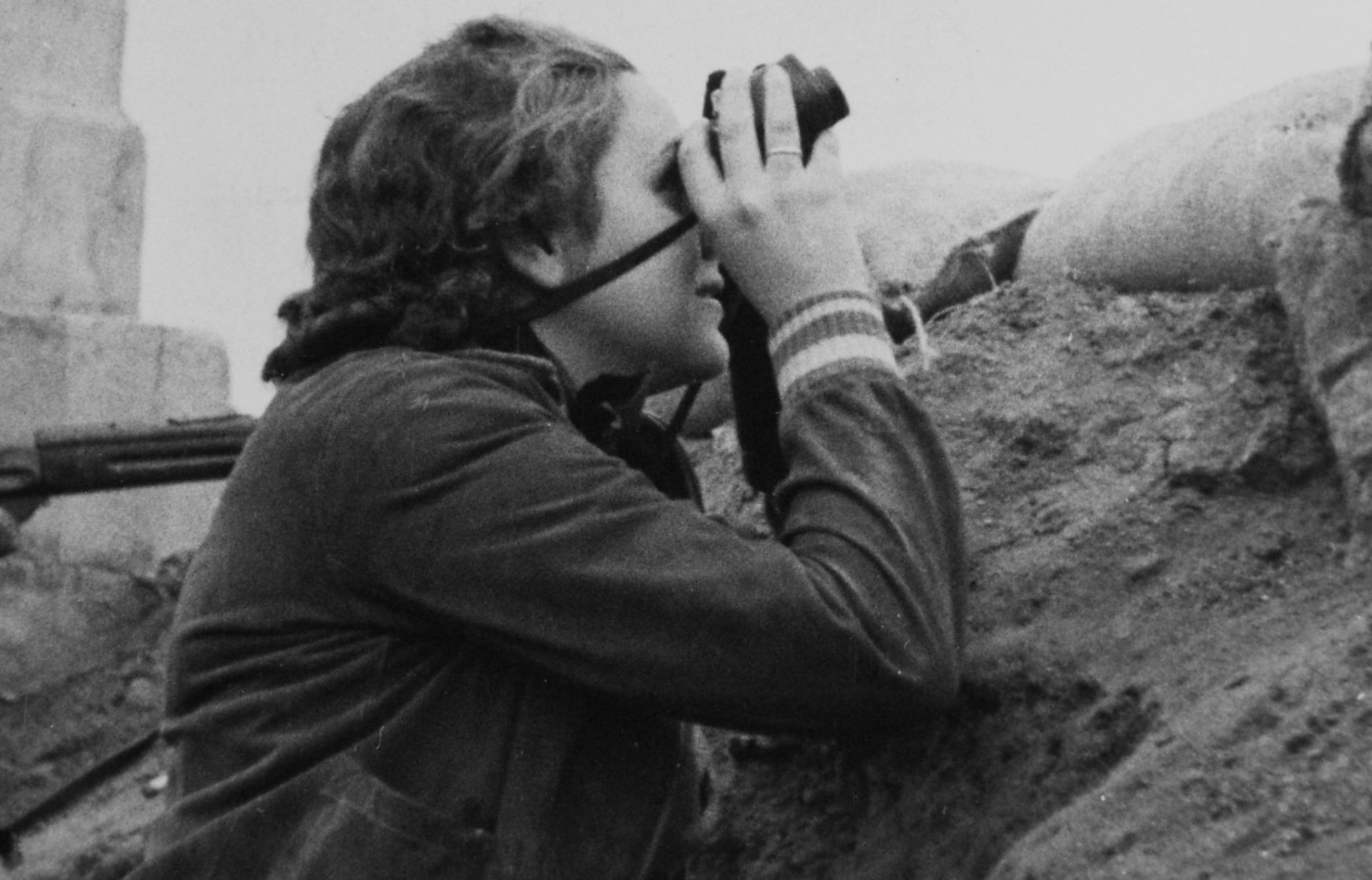 Black and white photo of a woman in a bunker looking toward the sky with binoculars