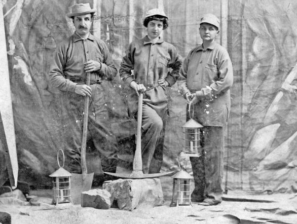 Black and white photograph of three people dressed in miners' clothing holding mining tools