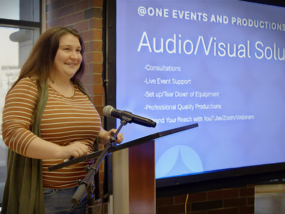 A woman stands at a podium with a presentation on a screen behind her. Some words are visible on the projection screen, including "audio/visual solutions".