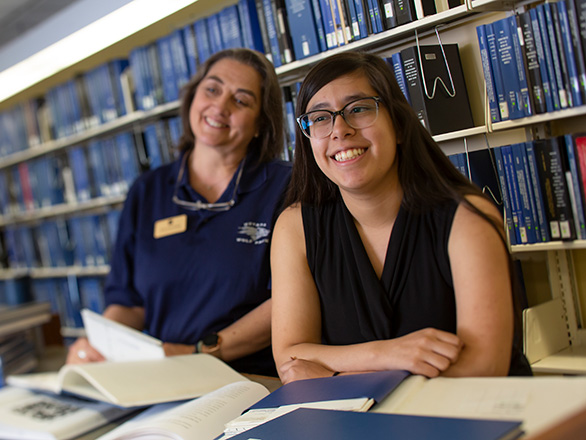 Two women stand in front of the thesis wall, filled with blue-spined booked.