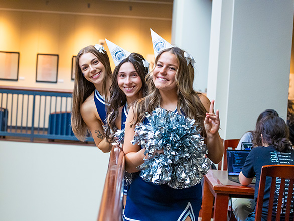 Three cheerleaders pose in the Knowledge Center. Two wear festive sesquicentennial party hats.