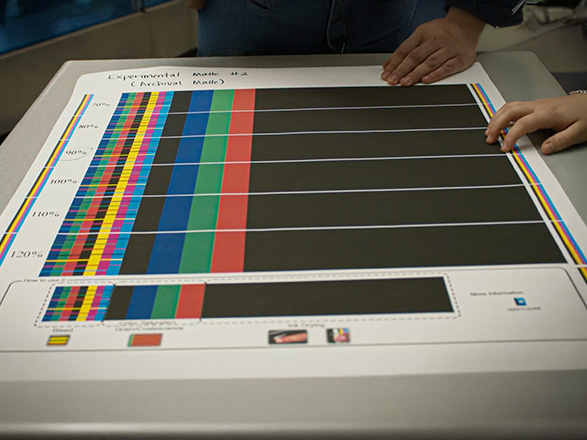 Color calibration test chart with vertical color bars and percentage markings laid flat on a table, with two people’s hands visible at the edges.