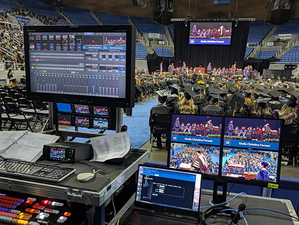 View from a video production control station with monitors, audio mixer, and laptop in the foreground, overlooking a large indoor graduation ceremony where graduates in caps and gowns face a stage in an arena.