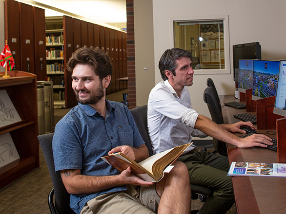 Two men sit at a row of computers in the Basque Library. One pages through a book.