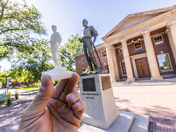A small, 3D printed statue is held up in front of the original, large statue outside of the DeLaMare Library.