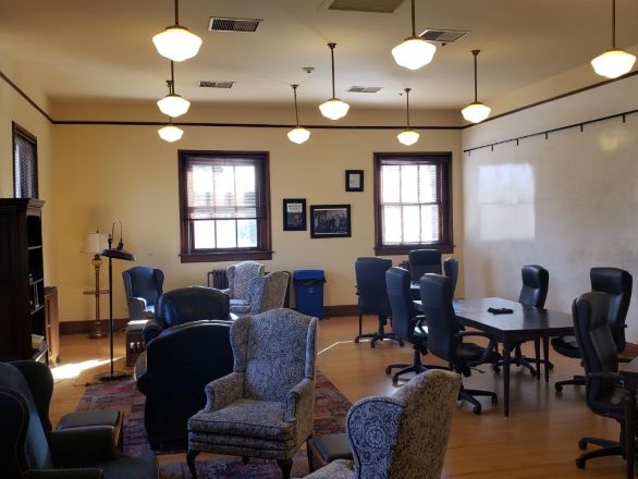 The Lilli Brant room with a large wall-sized white board, comfortable chairs, a bookshelf, two windows, and a meeting table.