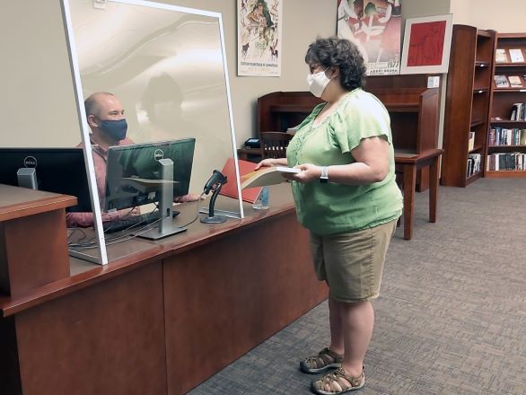 Man wearing face covering sitting at a library desl behind a barrier. He is helping a woman holding a book who is also wearing a face covering.