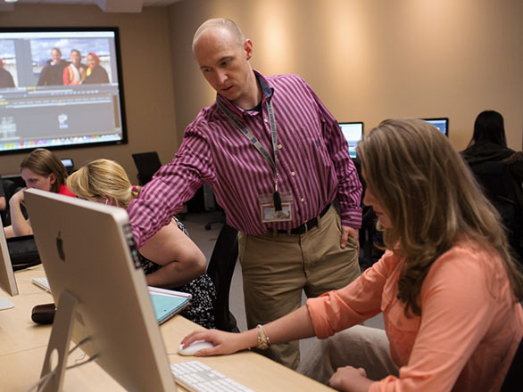 Students sitting at computer stations with instructor pointing to a student's computer monitor