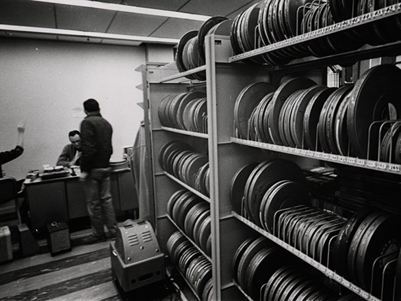 This view shows a man at a service desk and a patron with his back to camera with shelves of reel-to-reel films in the foreground. ID: UNRA-P2062-05