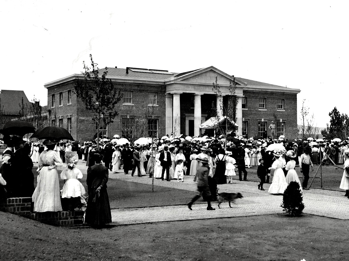 This scene shows the Mackay Statue unveiling during the dedication ceremony for the Mackay School of Mines with a crowd of dressed-up spectators milling around the Quadrangle in the foreground. 