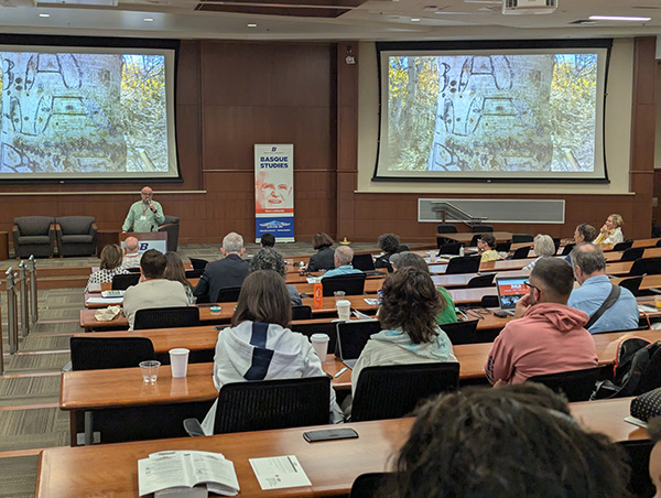 Inaki stands at the front of a classroom at a podium. People sit in rows facing him. There are pictures of arborglyphs on screens at the front of the room. A poster at the front reads: "Basque Studies".