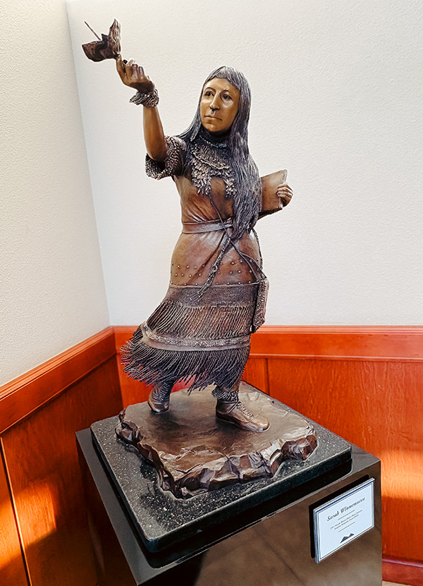 Bronze sculpture of a woman wearing a flowing dress holding a book in one hand with a bird perched on her other hand.