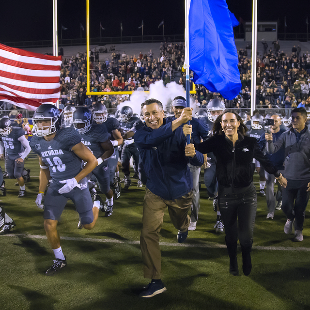 University president Brian Sandoval runs across a football field with the football team, holding a flag and smiling.
